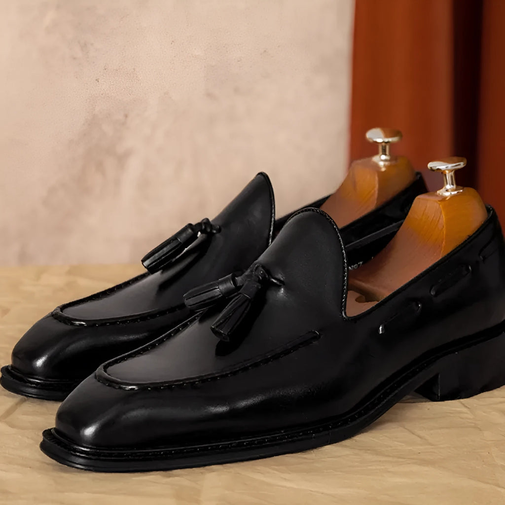 Men's black leather slip-on loafers with fringe detailing on a wooden floor