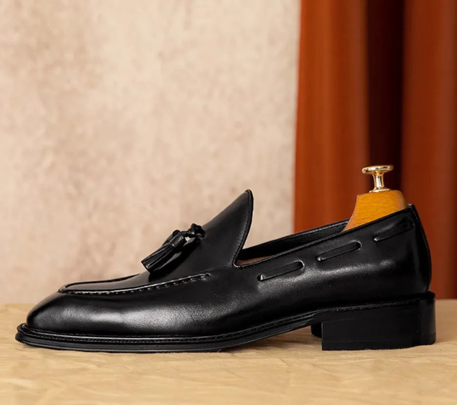Men's black leather slip-on loafers with fringe detailing on a wooden floor