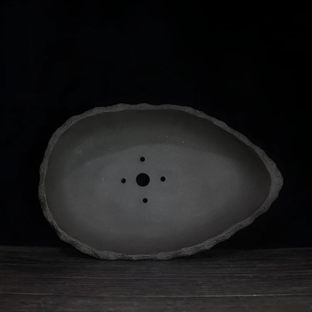 Handmade purple clay flowerpot in a unique shape, shown on floor beside a plant.