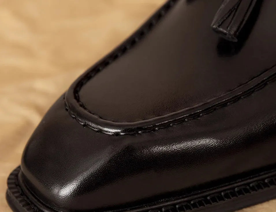 Men's black leather slip-on loafers with fringe detailing on a wooden floor