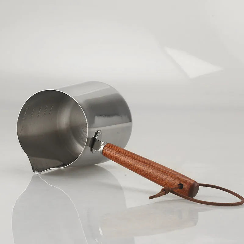 Stainless steel barista milk cup with measurement scale markings and a wooden handle.
