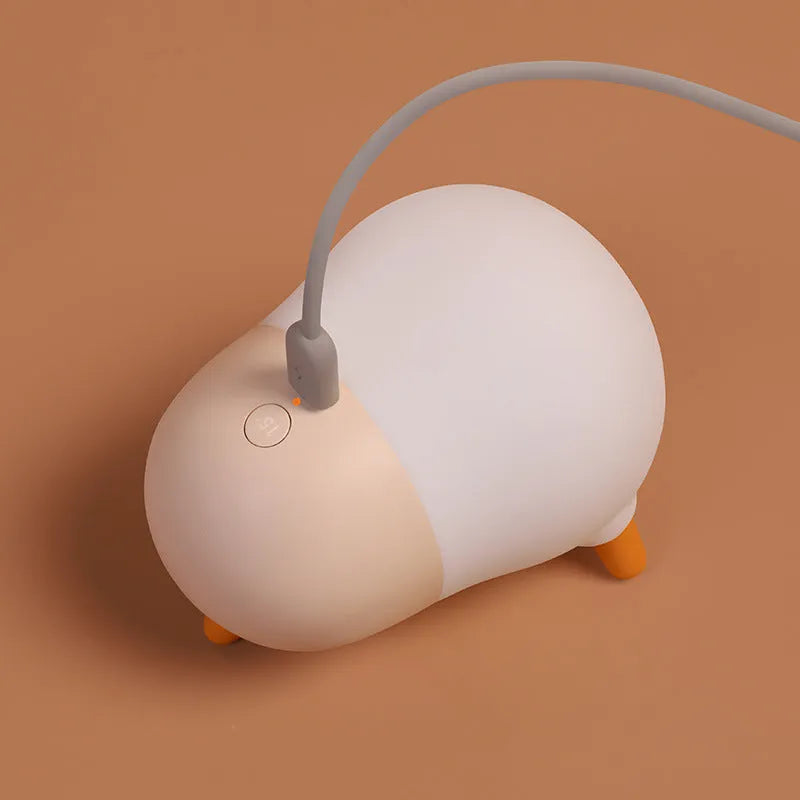 Pecking rice chicken small night light with warm yellow glow on a wooden bedside table, USB charging cable visible