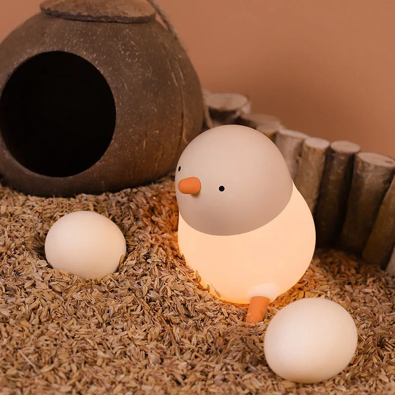 Pecking rice chicken small night light with warm yellow glow on a wooden bedside table, USB charging cable visible