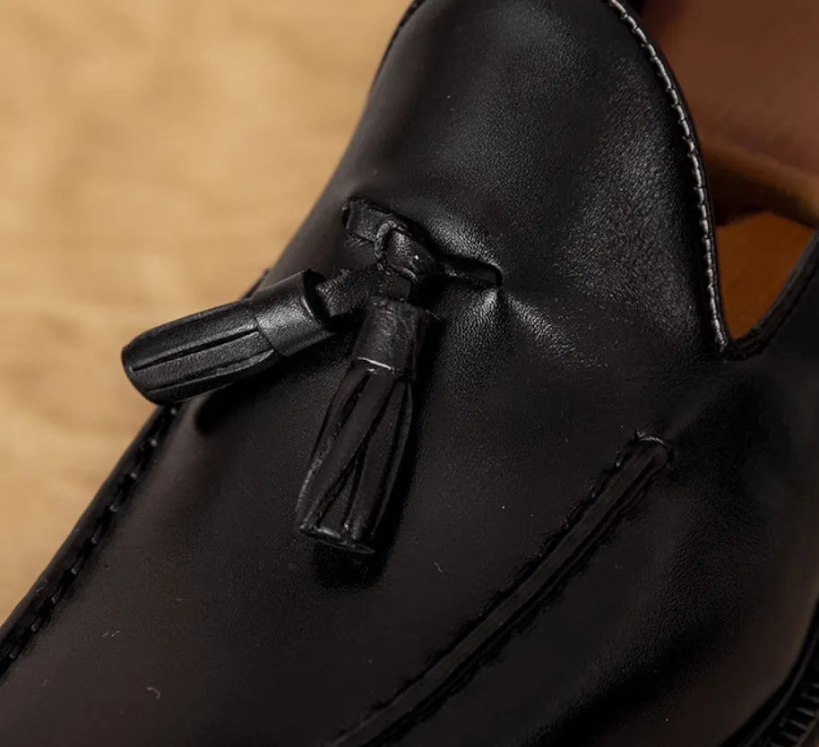 Men's black leather slip-on loafers with fringe detailing on a wooden floor