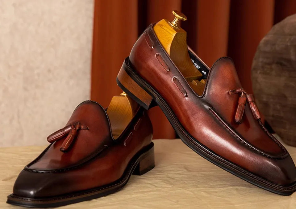 Men's black leather slip-on loafers with fringe detailing on a wooden floor