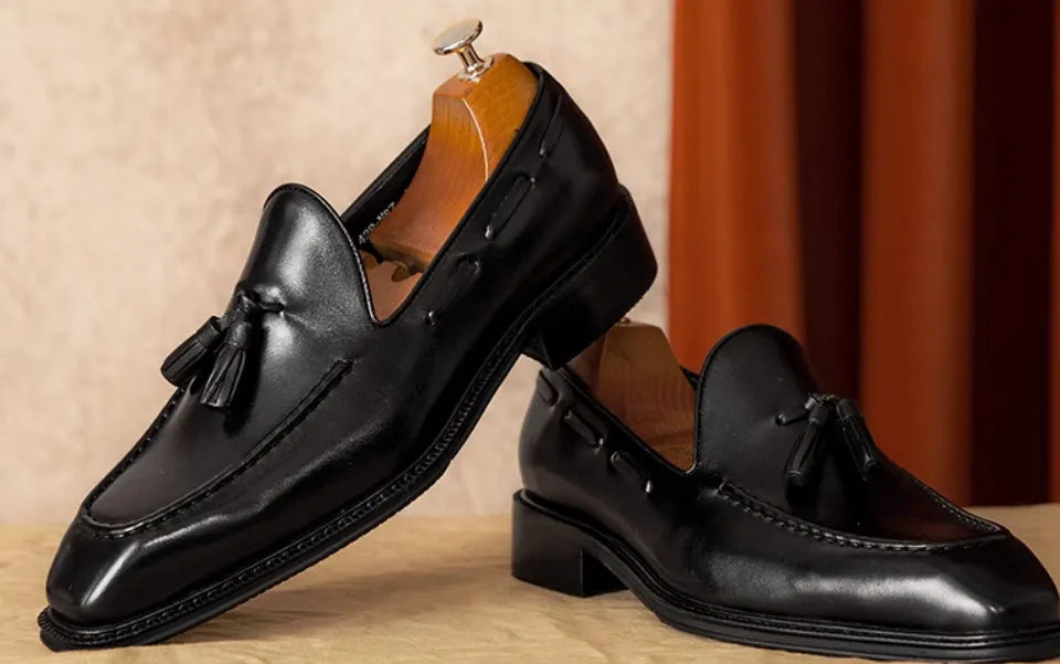 Men's black leather slip-on loafers with fringe detailing on a wooden floor